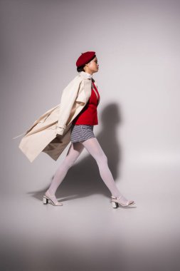 Side view of stylish asian woman in trench coat walking on grey background with shadow 