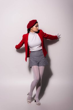 Full length of trendy asian model in red beret and jacket touching grey background 