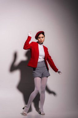 Full length of stylish asian model in red beret and jacket standing on grey background with shadow 