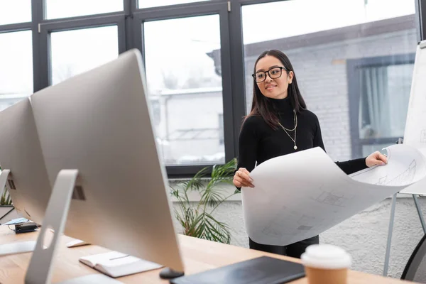 Stock image Smiling asian interior designer holding blueprint and looking at computer monitor in studio 