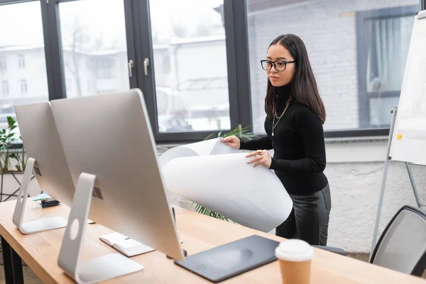 stock image Asian interior designer holding blueprint near computers in office 