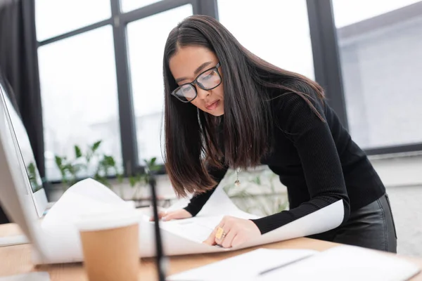 stock image Young asian designer in eyeglasses looking at blueprint near computers in studio 