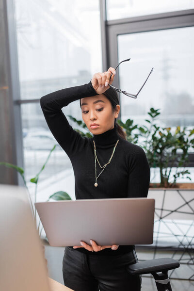 Беспокойный азиатский дизайнер интерьеров держит очки и ноутбук в студии 