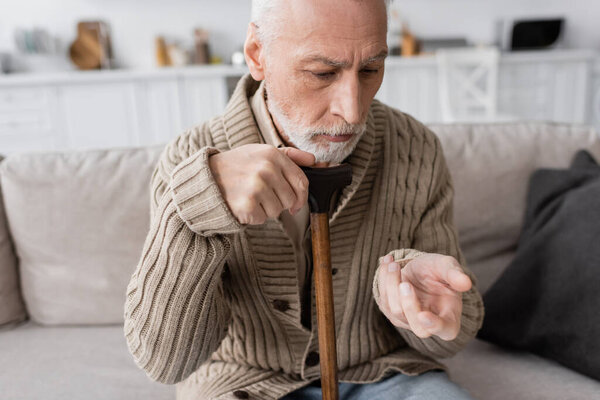 senior man in knitted cardigan sitting with walking cane and looking at trembling hand while suffering from parkinson disease