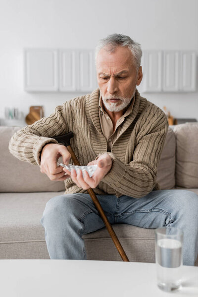 aged man with parkinson syndrome holding pills in trembling hands while sitting on couch at home