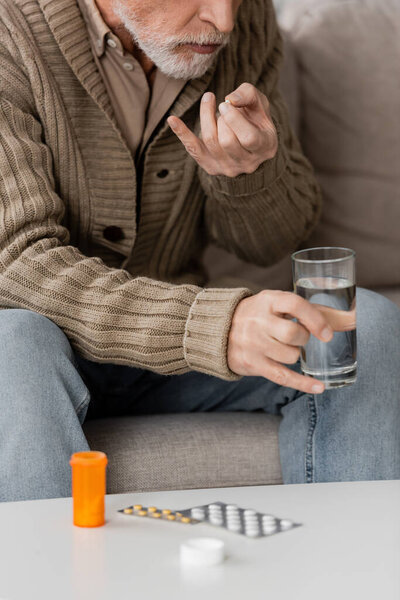 cropped view of man with parkinson disease holding glass of water near pills on table at home