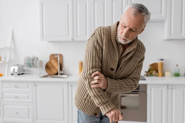 grey haired man suffering from parkinsonism and hands tremor standing with walking cane in kitchen at home