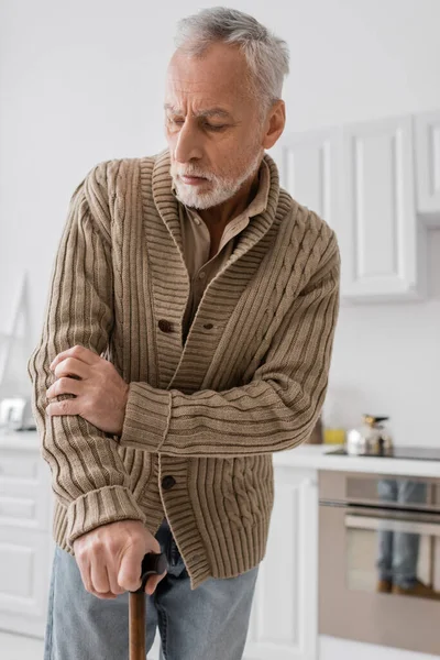 tensed man in knitted cardigan standing with walking cane in kitchen while suffering from parkinson disease