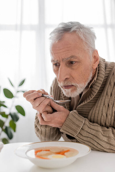 depressed man with parkinsonian syndrome sitting with spoon in trembling hands near soup in kitchen