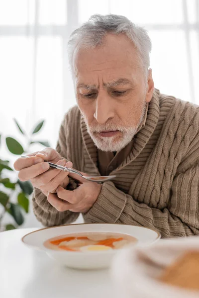 stock image senior man with parkinson disease holding spoon in trembling hands near plate with soup in kitchen 