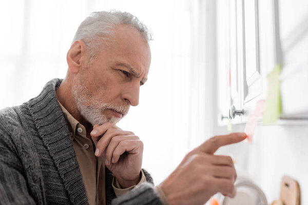 side view of pensive man with alzheimer syndrome touching chin and pointing at blurred sticky note in kitchen