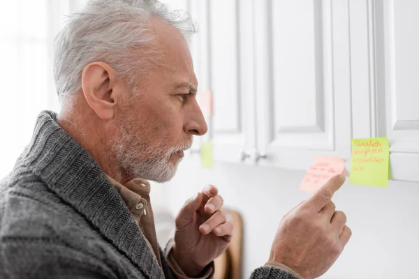 side view of thoughtful man with alzheimer syndrome pointing at blurred sticky note in kitchen
