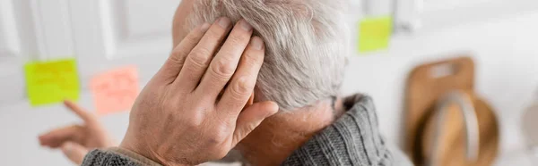 stock image aged man with alzheimer disease touching head and pointing at blurred sticky notes in kitchen, banner