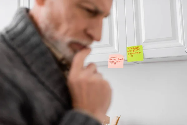 blurred thoughtful man with memory loss touching chin while thinking near sticky notes in kitchen