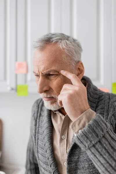 tensed man suffering from memory loss and touching head while thinking near blurred sticky notes in kitchen
