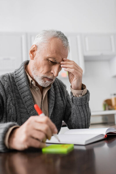 thoughtful man touching forehead while sitting with felt pen near sticky notes and empty notebook