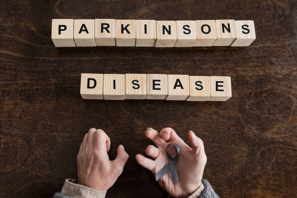 top view of cropped man holding grey ribbon in trembling hands near blocks with parkinsons disease inscription on wooden surface