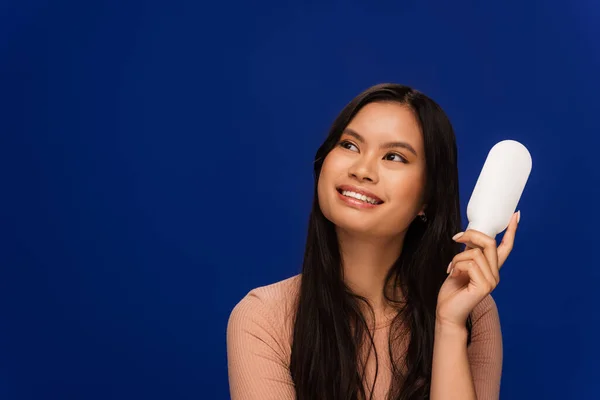 stock image Cheerful asian model holding body lotion and looking away isolated on blue 