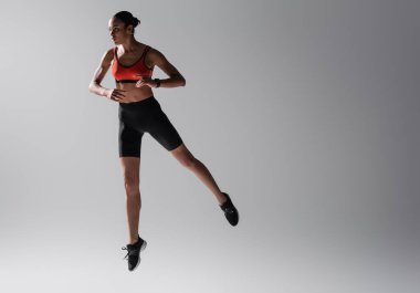 full length of tattooed african american woman in sports bra and shorts jumping on grey background 
