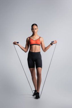 full length of sportive african american woman standing with jumping rope on grey 