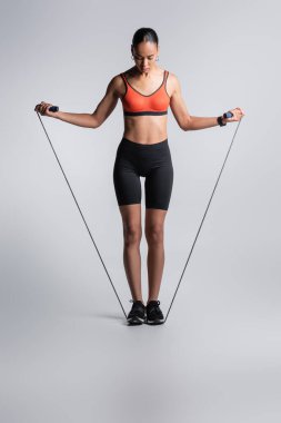 full length of sportive african american woman standing with skipping rope on grey 