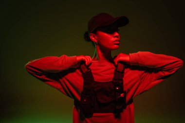 african american woman in sweatshirt and vest posing on green with red light 