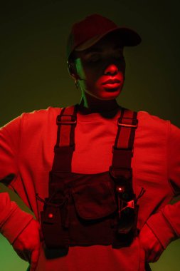 african american woman in sweatshirt and vest posing with hands on hips on green with red light 