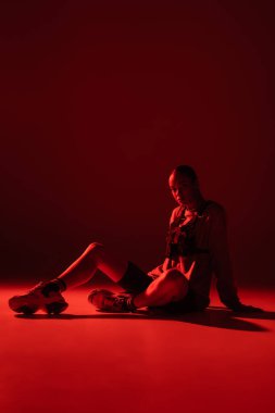 full length of young african american woman in bike shorts and sweatshirt sitting on dark red background 