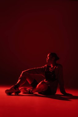 full length of young african american woman in bike shorts and sweatshirt sitting on dark red background 