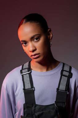 stylish african american woman in sweatshirt and vest looking at camera on grey 