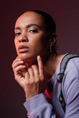 young african american woman in violet sweatshirt and vest looking at camera on dark purple 