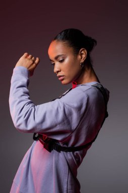 brunette african american woman in sweatshirt and vest posing on grey and purple 