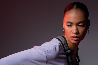 brunette african american woman in sweatshirt and vest looking at camera on grey and purple background 