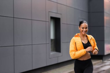 positive african american sportswoman in yellow puffer jacket jogging outside 