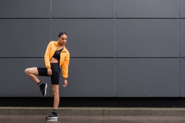 full length of brunette african american sportswoman in bike shorts and yellow puffer jacket posing outside 