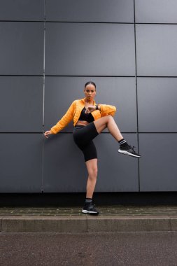 full length of stylish african american sportswoman in bike shorts and yellow puffer jacket exercising outside 
