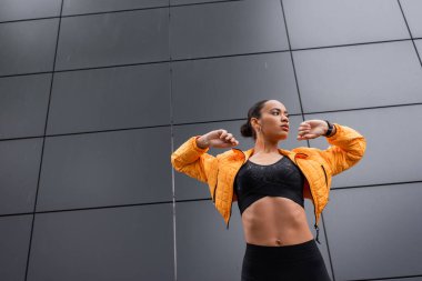 low angle view of brunette african american sportswoman in sports bra and yellow puffer jacket exercising outside 