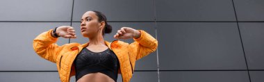 low angle view of brunette african american sportswoman in sports bra and yellow puffer jacket exercising outside, banner 