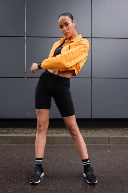 full length of stylish african american sportswoman in bike shorts and yellow puffer jacket posing near grey building 