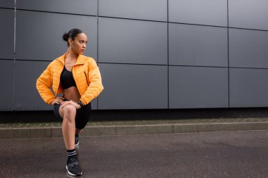 full length of stylish african american sportswoman in bike shorts and yellow puffer jacket working out near grey building 