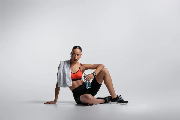 young african american woman in sports bra and bike shorts sitting with sports bottle and towel on grey background