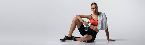 young african american woman in sports bra and bike shorts sitting with sports bottle and towel on grey background, banner 