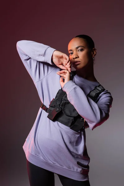 stock image young african american woman in sweatshirt and vest looking at camera while posing on grey and purple 