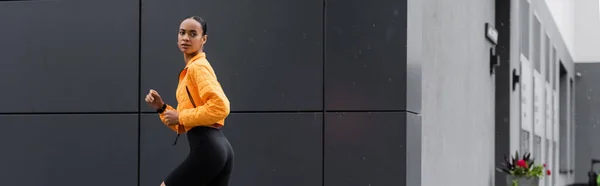 Stock image brunette african american sportswoman in bike shorts and yellow puffer jacket running outside, banner 