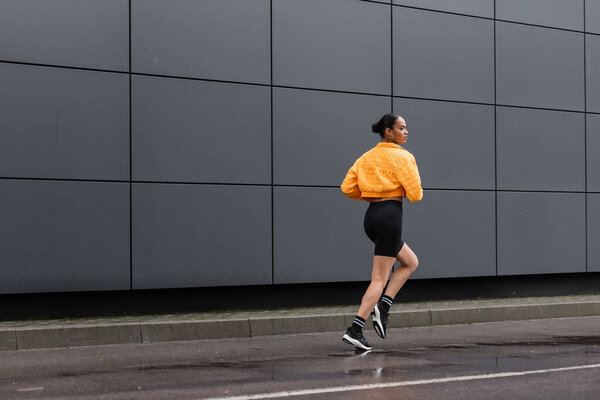 full length of brunette african american sportswoman in bike shorts and yellow puffer jacket jogging outside 