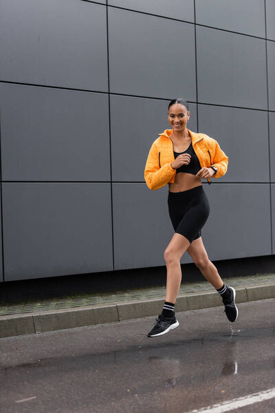 full length of cheerful african american sportswoman in bike shorts and yellow puffer jacket exercising outside 