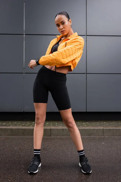 Stock image full length of stylish african american sportswoman in bike shorts and yellow puffer jacket posing near grey building 