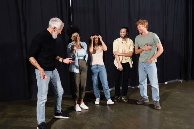 bearded teacher of acting skills talking to cheerful interracial students in theater school