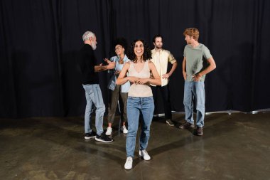 smiling multiracial woman rehearsing near actors and art director talking on stage in theater 