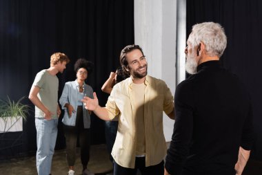 young and happy artist talking to acting skills teacher near interracial student in theater school